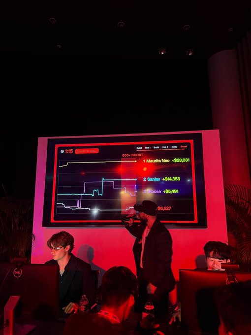A presenter stands before a large screen displaying a live "600+ BOOST" trading leaderboard with tickers like "Maurits Neo," "Sunjay," "Mooso," and "Neo" showing gains of +$28,031, +$14,353, +$5,491, and +$5,527 respectively, while seated competitors work at monitors during the Frontier and Solana-powered competition. The screen's bullish red glow and competitive leaderboard visuals capture the energetic atmosphere of the trading event highlighted in the caption.