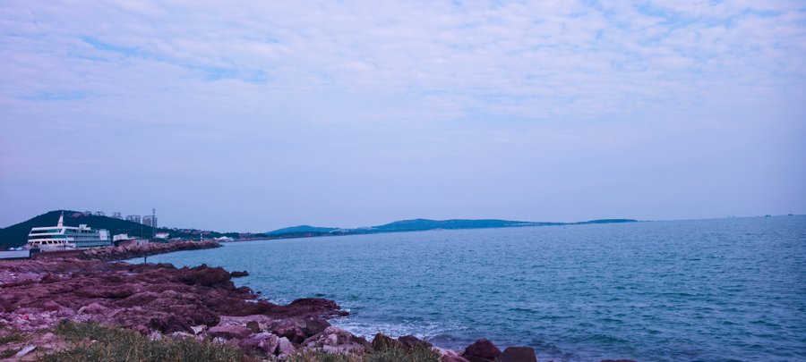 A serene coastal landscape features a rocky shoreline with purple-tinged rocks, calm blue water, distant hills, and a partly cloudy sky.