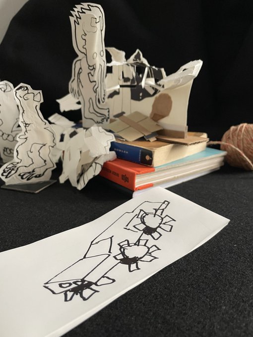 Black and white paper cutouts of cartoon characters standing on and around a stack of books, with a ball of twine nearby and a hand-drawn sketch in the foreground.