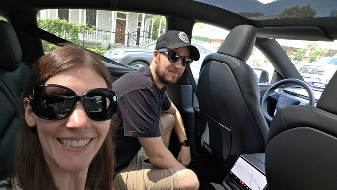 A smiling woman wearing sunglasses and a bearded man in a baseball cap sit inside a modern, sleek vehicle with a large touchscreen display, capturing a selfie to commemorate their first ride in a robotaxi. The couple's excited expressions match the futuristic experience of riding in the autonomous vehicle.