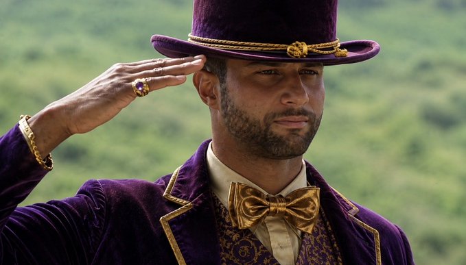 A man wearing an ornate purple velvet suit with gold accents and a matching top hat, tipping his hat while wearing a ring with a large purple stone.