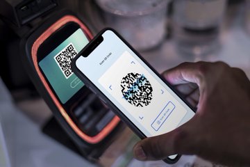A smartphone displays a QR code scanning interface being aimed at another QR code on a handheld device, illustrating a digital payment transaction. The image aligns with the post about the Gujarat government's initiative to implement Central Bank Digital Currency (CBDC) for National Food Security Act (NFSA) beneficiaries, showcasing the QR-based technology enabling this digital payment innovation.