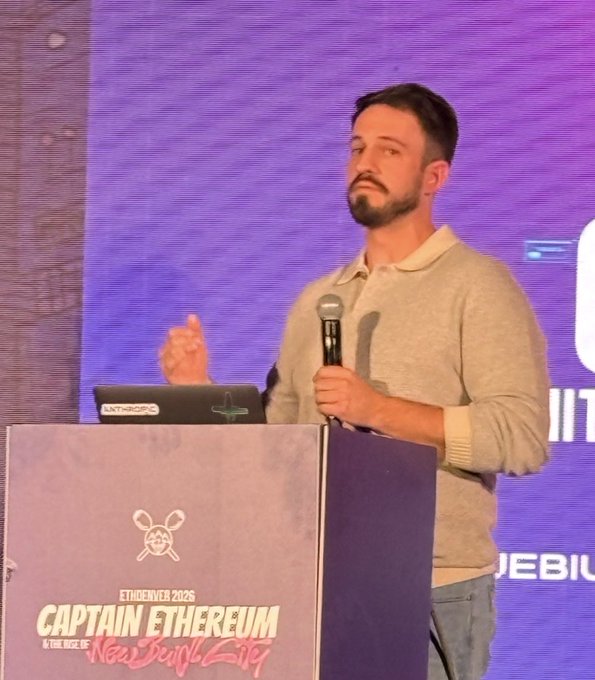 A bearded man in a beige sweater speaks at the EthDenver 2026 'Captain Ethereum' podium, holding a microphone with an Anthropic-branded laptop nearby.