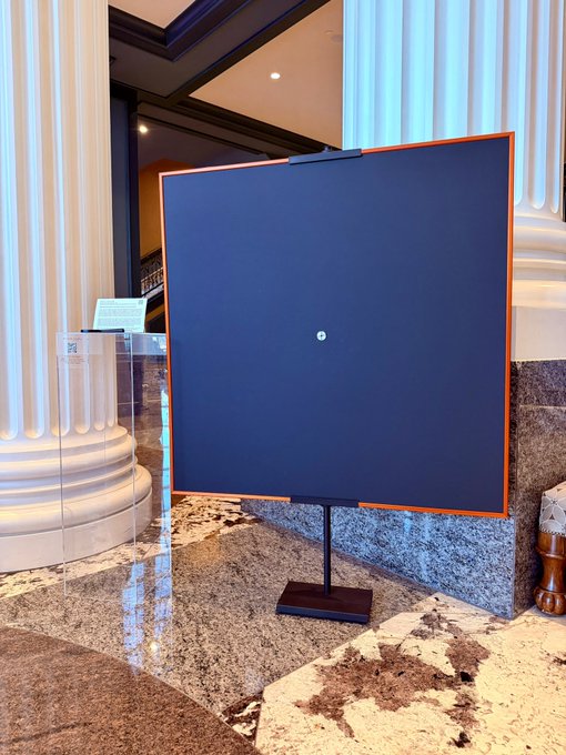 A dark blue display panel with an orange border stands on a black pedestal in a hotel lobby with marble floors and white columns.