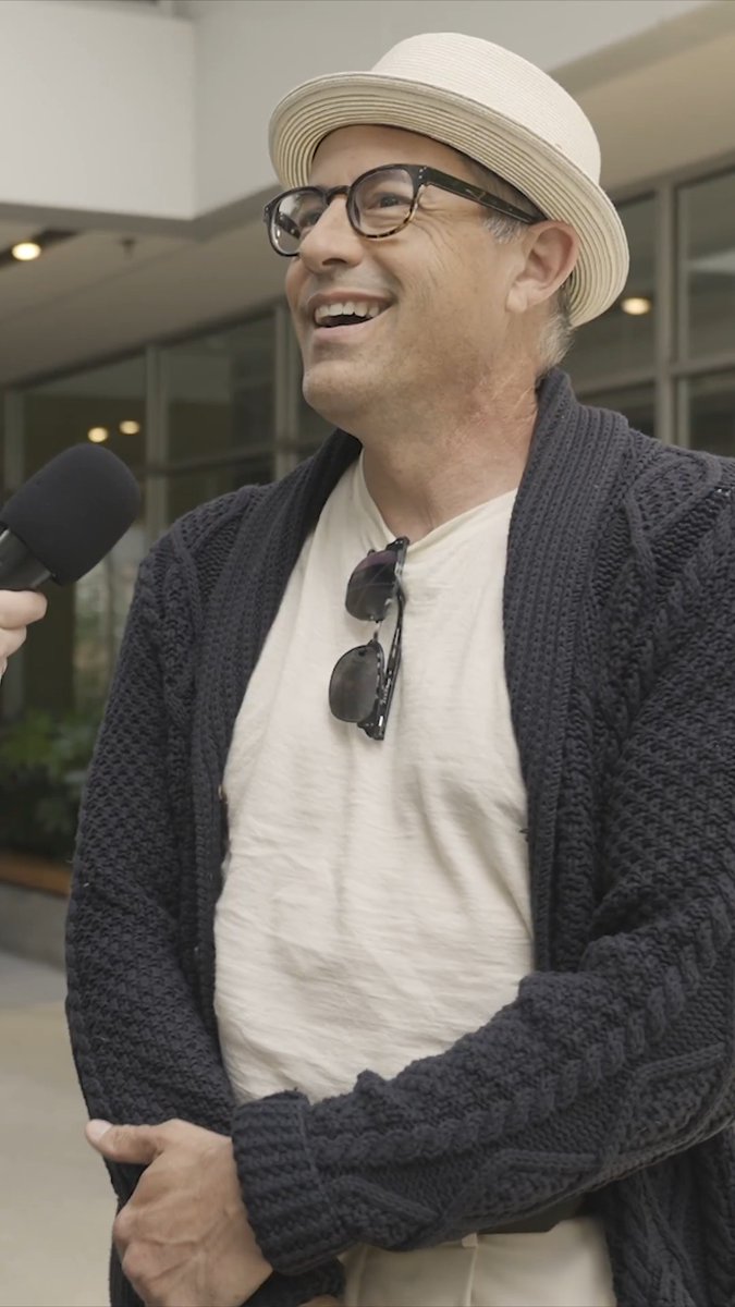 A smiling man wearing a cream hat, glasses, cream t-shirt, and dark knit cardigan is being interviewed with a microphone.