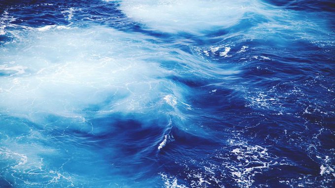 An abstract close-up image of swirling ocean waves in deep blue and white foam patterns.