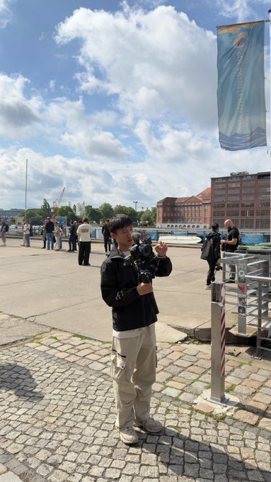 A young man in a black jacket and beige cargo pants stands on a cobblestone plaza holding a camera, preparing to film content. The banner overhead reads "EDWARDS." The scene reflects the post's theme of moving from anonymous online interactions to in-person content creation, capturing the creator behind the lens at work in a public setting.