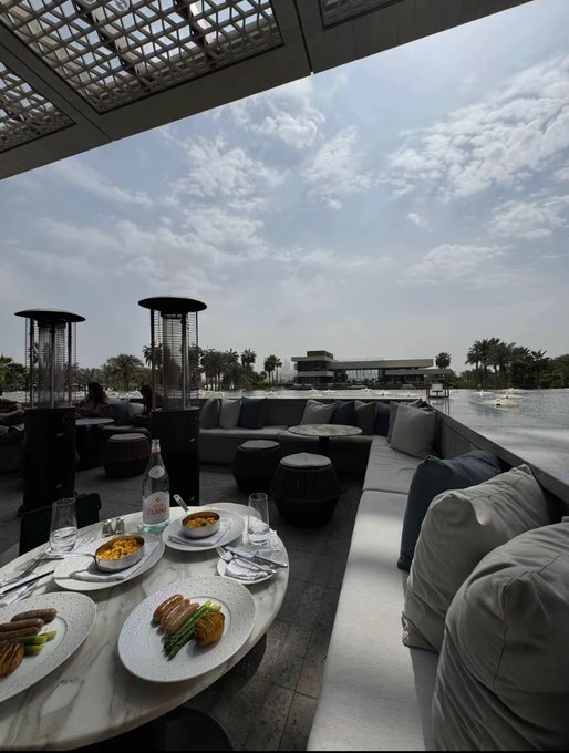 A luxury outdoor lounge overlooking a waterfront with palm trees and a modern building in the background, featuring a marble table set with plated meals and a bottle. The serene, upscale setting complements the upbeat "GM CT" morning greeting, suggesting a refined start to the day.