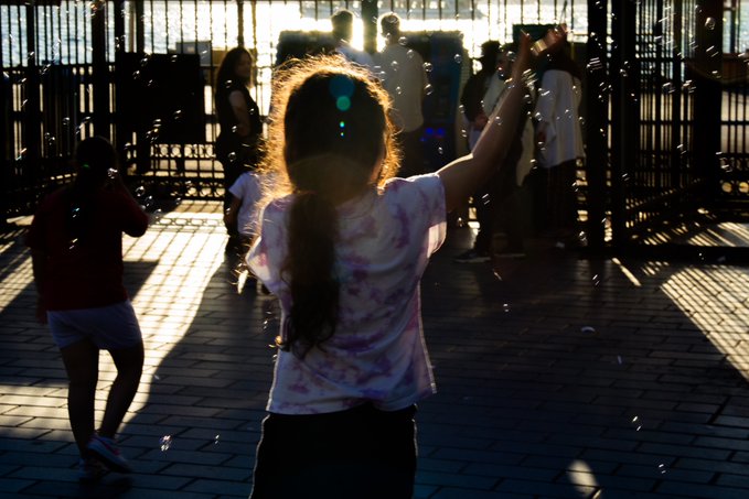 A photograph captures a girl from behind reaching toward floating bubbles, silhouetted against bright sunlight streaming through a gate.