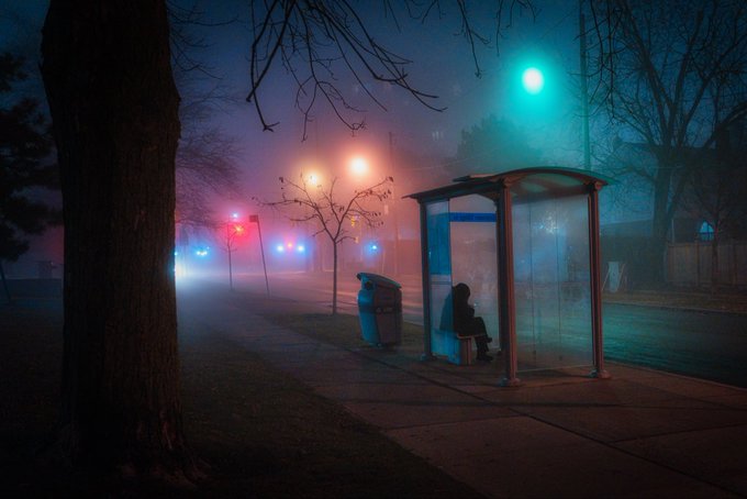 A moody nighttime photograph of a lone figure sitting inside a glass bus shelter on a foggy street, illuminated by a cyan streetlight and distant red and blue lights.
