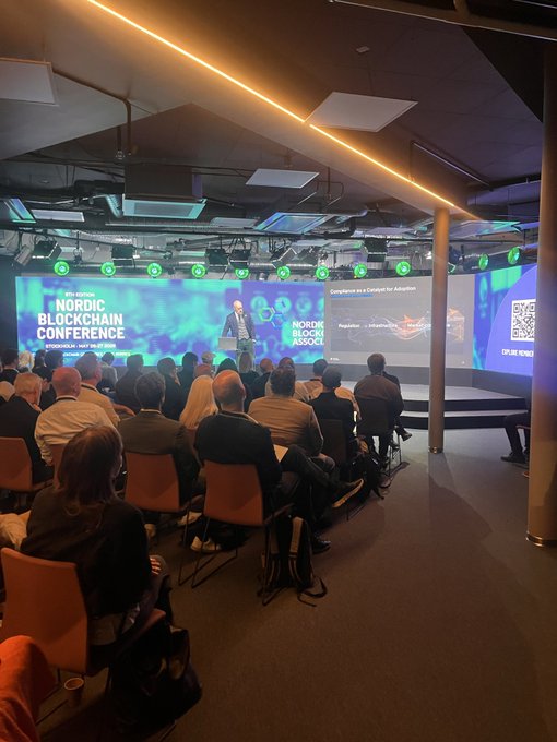 The image shows a large group of people sitting down, possibly attending a conference or event related to blockchain technology. A man is standing up and giving a presentation at this gathering. The attendees are engaged with the speaker's words as they listen intently. In the background, there is a screen displaying information about the QROMO platform, which is an open-source blockchain solution for Ethereum. The image also includes a text that reads "Today QROMO went live at the Nordic Blockchain Conference People scanned. Engaged.received instant Solana rewards. Real-world blockchain engagement is here."