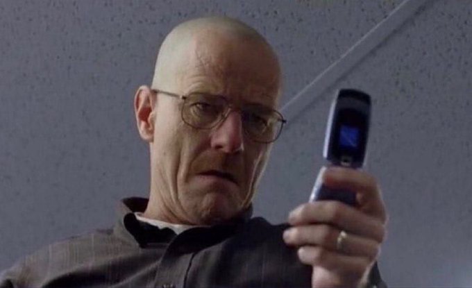 The character Walter White from Breaking Bad holding up an old flip phone while wearing glasses.