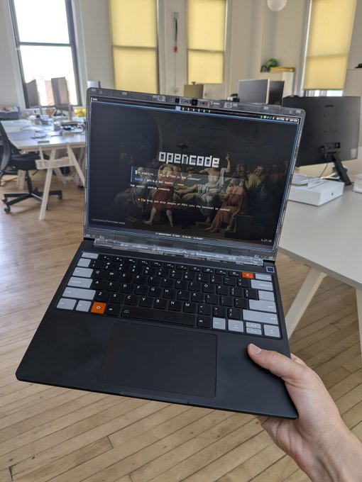 A hand holds up a laptop displaying the "opencode" website in what appears to be a modern office space with large windows and wooden floors, fitting the "ferryman.io office for today" caption. The setting suggests a casual workday environment at the ferryman.io office, with the device showcasing the company's open-source coding platform.