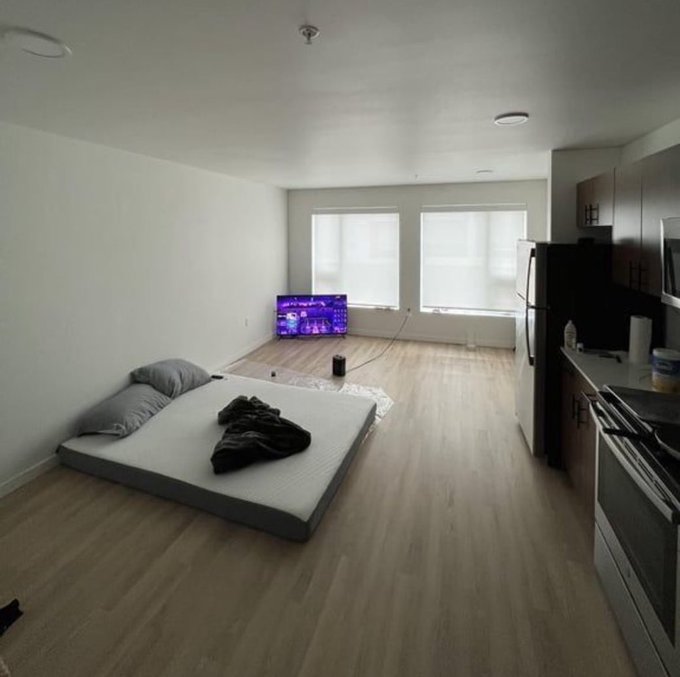 An interior view of an unfurnished studio apartment featuring a mattress on the floor, a TV, wood-look flooring, and a kitchen area.