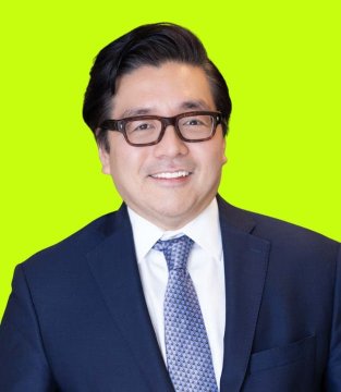 A professional headshot of a smiling man with dark hair wearing black-rimmed glasses, a navy blue suit, white shirt, and blue patterned tie against a bright green background.