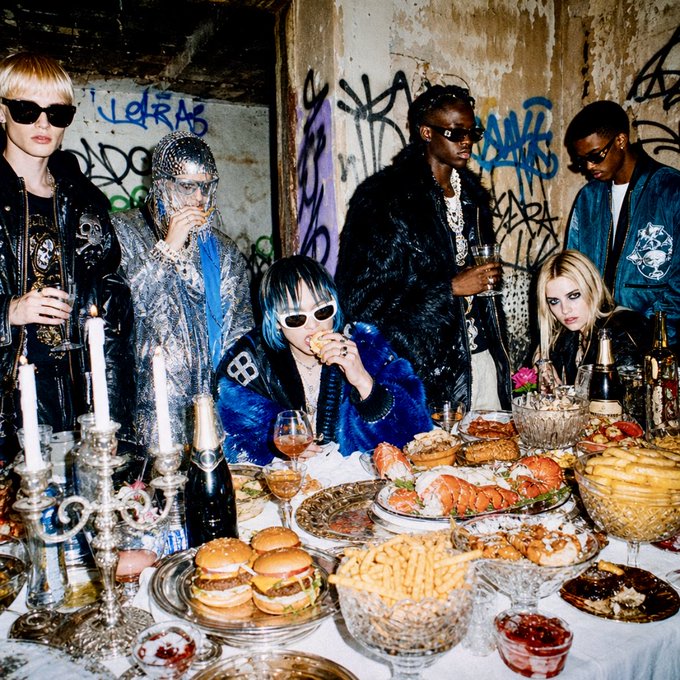 A group of seven stylishly dressed people, including a blonde woman in sunglasses and a person with blue hair, gathered around a lavishly laid banquet table with sliders, fries, and champagne.