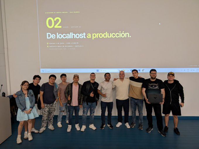 The image features a group of people standing next to each other, posing for a photo. They are all wearing casual clothing, with some individuals dressed more formally than others. The group consists of at least nine people, including both men and women. One person in the group is holding a laptop computer. In front of them, there is a large screen displaying text that reads "02 De Locallos A Productividad", which translates to "02 Local Host Production". This suggests that they are gathered for an event related to local production or a similar project.