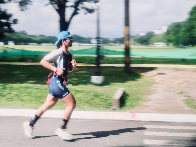An athlete in a triathlon outfit runs along a road, wearing a blue cap, sunglasses, and a race bib, with motion blur showing her movement through a green, tree-lined area.