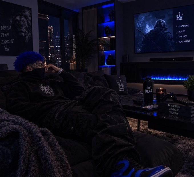 A person with blue hair relaxes under a black blanket in a dimly lit modern living room, with a TV displaying 'DISCIPLINE, BUILDS, THE LIFE YOU WANT' and a lion tapestry reading 'DREAM PLAN EXECUTE'.
