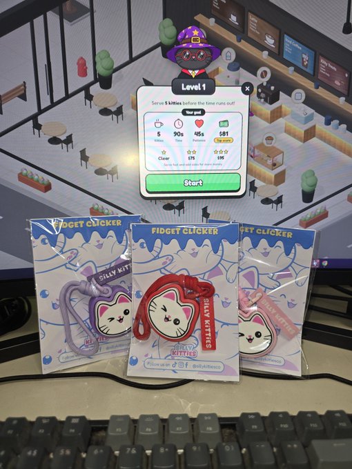 Three 'Fidget Clicker - Silly Kitties' toys in purple, red, and pink packaging sit on a desk in front of a monitor displaying a Level 1 café management game by BulkATeaco.