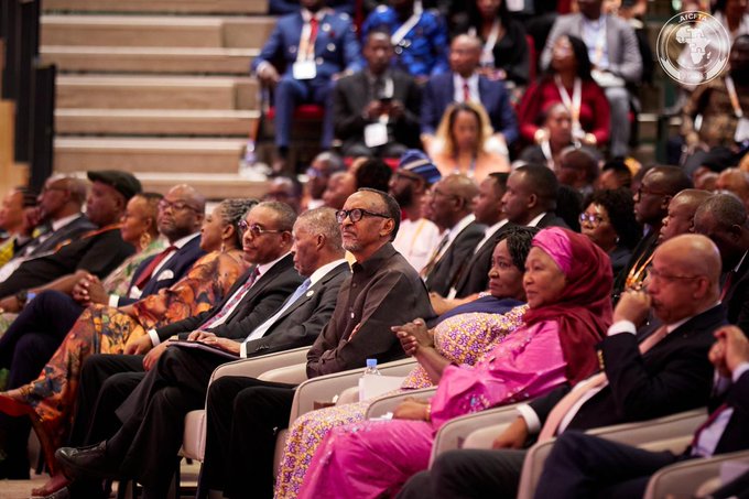A large group of formally dressed attendees, including dignitaries in traditional African attire and suits, seated in an auditorium with an AU (African Union) emblem visible in the corner.