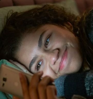 A close-up photo of a young woman with curly hair smiling softly while lying down and holding a rose gold smartphone, appearing to take a selfie.