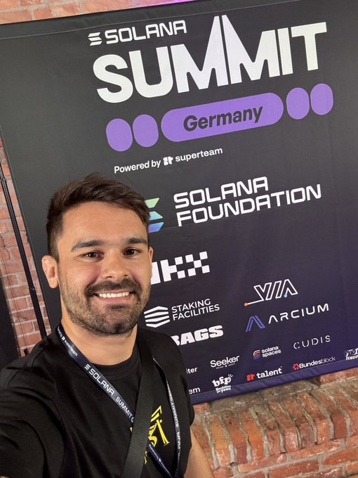 A man smiling at the camera in front of a Solana Summit Germany banner powered by Superteam, with various sponsor logos including Solana Foundation, OKX, and others visible.