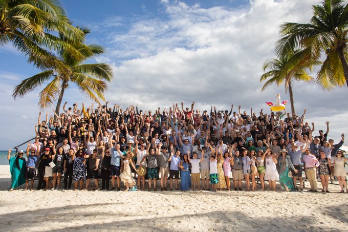 A large group of people posing together on a tropical sandy beach between palm trees, cheering with raised arms under a partly cloudy sky, with ocean visible in the background.