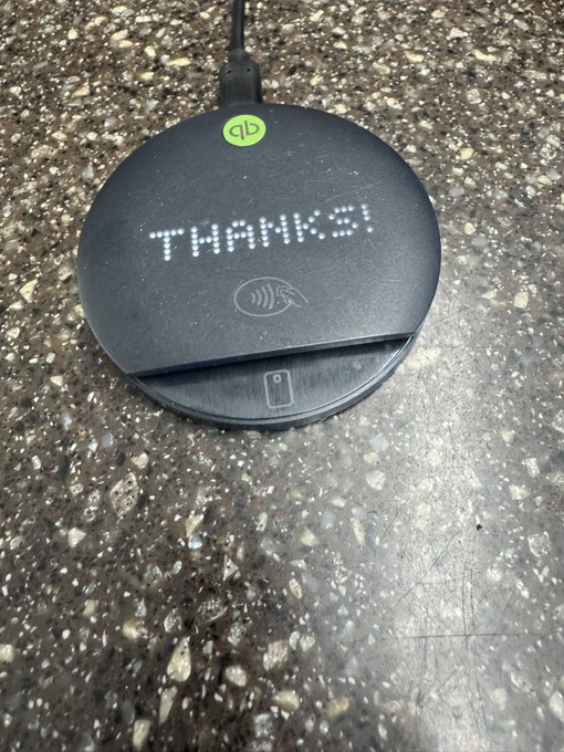 A black circular wireless charging pad with a green "qb" logo sits on a speckled countertop, displaying "THANKS!" in white LED text with a contactless payment symbol.