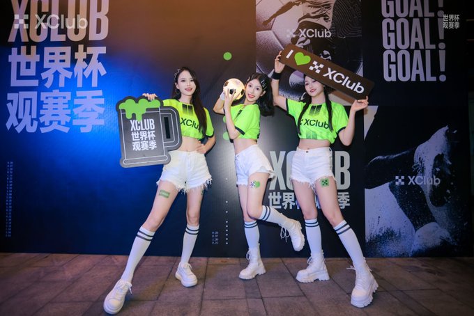 Three young women in green and white XClub promotional outfits posing with signs at what appears to be a World Cup viewing event ("世界杯观赛季" / World Cup Watch Party).