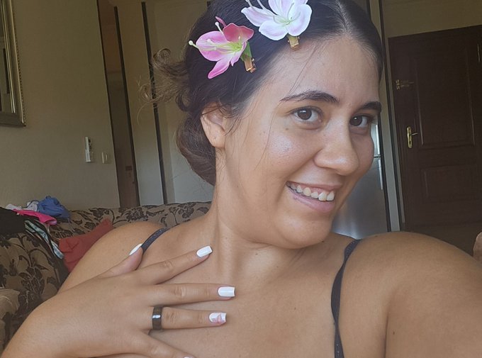 A selfie of a smiling young woman with dark hair adorned with pink and white flowers, wearing a ring and sporting white nail polish with a small design.