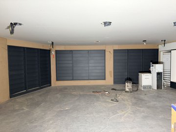 An empty garage or workshop interior with tan walls, dark blue/black louvered panels, and concrete floor, with some equipment and cables scattered near the back wall.