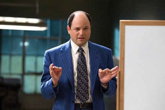 A still image of George Costanza from Seinfeld, played by Jason Alexander, wearing a blue pinstripe suit and patterned tie, gesturing with both hands while speaking in what appears to be an office setting.