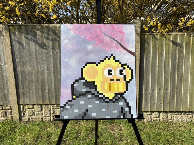 A pixel art painting on a black easel outdoors depicts a yellow character with large eyes emerging from a gray rock, set against a background of a pink tree and sky, positioned in front of a wooden fence.