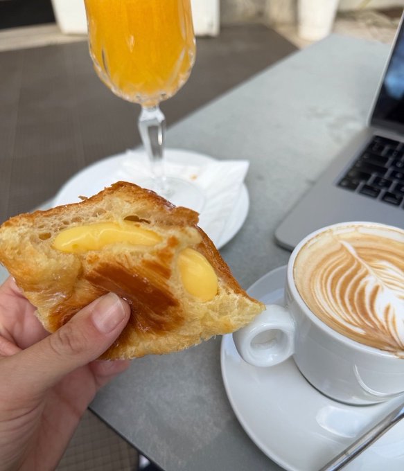 A hand holds up a flaky pastry filled with creamy yellow custard, paired with a beautifully latte-art cappuccino and a glass of orange juice at a café table beside a MacBook. The image playfully illustrates the post's humor—contrasting a wholesome, civilized breakfast against the carnivorous chaos of "eating a steak for breakfast."