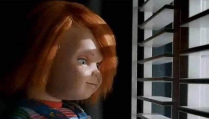 A still from a live-action film showing the character Chucky peeking through window blinds.