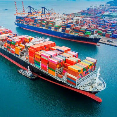 An aerial view of a large container ship docked at a busy port, loaded with multicolored shipping containers, with a smaller tugboat nearby.