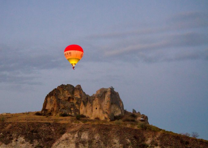 A red and yellow hot air balloon floats above eroded rock formations and a dry hillside during golden hour.