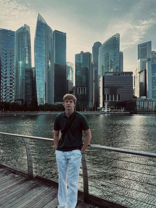 A young man poses confidently on a waterfront promenade with Singapore's iconic skyline—including the HSBC tower—in the background, ready to connect at TOKEN2049 during Singapore Blockchain Week.