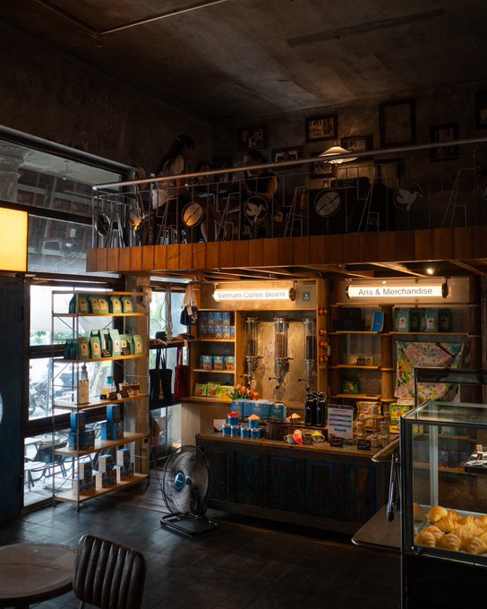 An interior photograph of a coffee shop or café featuring a mezzanine level lined with stools, a counter labeled 'Vietnam Coffee Beans' and 'Arts & Merchandise', and shelves stocked with coffee supplies and packaged goods.