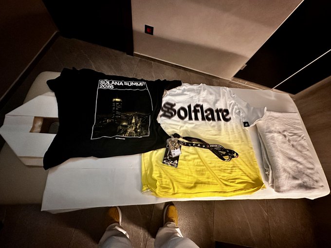 Two t-shirts laid out on a hotel bed—one black "Solana Summit 2026" shirt and one white-to-yellow gradient "Solflare" shirt—with the photographer's shoes visible at the bottom.