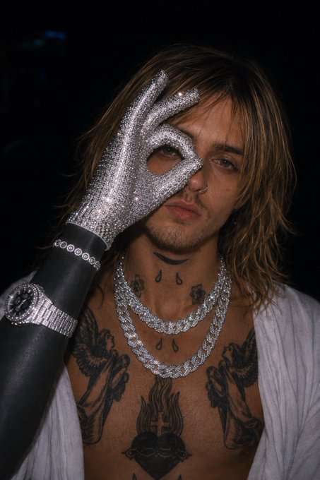 A young man with long hair and an array of tattoos, including angels, a sacred heart, and a teardrop design, holds up a sparkly silver glove while wearing multiple diamond chains and a large iced-out watch, exuding a glamorous yet edgy aesthetic that fits the "Back to business" theme.