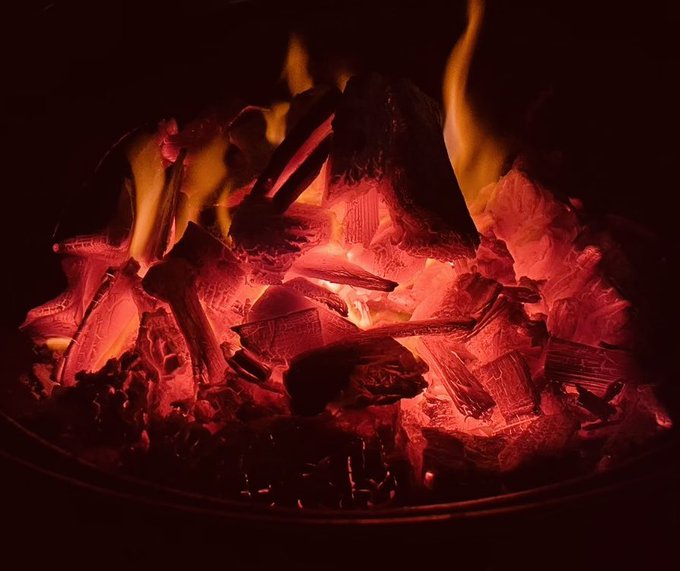 A close-up photograph of glowing red and orange embers and flames burning in a fire pit or grill against a dark background.