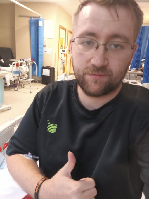 A selfie of a bearded man wearing glasses and a black t-shirt with a green logo, giving a thumbs-up while seated in what appears to be a medical or hospital room with blue privacy curtains and medical equipment in the background.