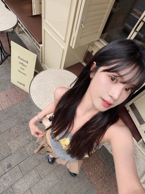 A high-angle selfie of a young woman with long dark hair wearing a yellow top, taken on a cobblestone street outside a café with a sign reading 'Wesh Brunch Coffee Dessert Dimas'.