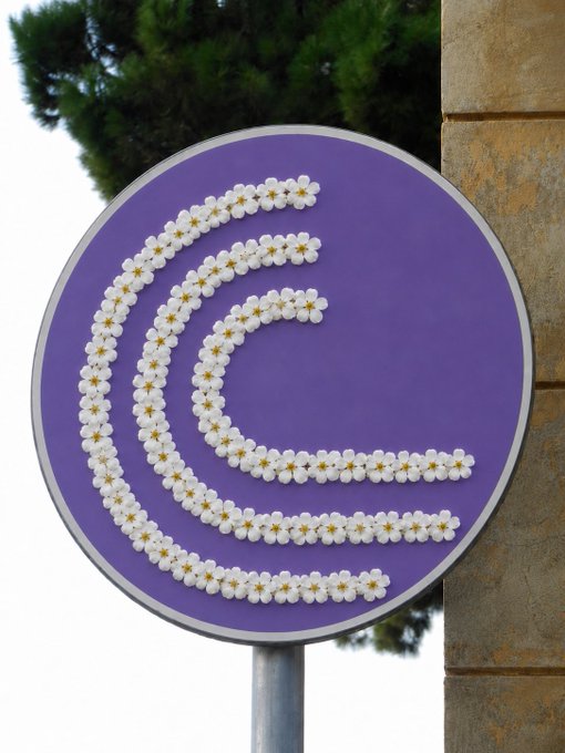 A round purple sign featuring a stylized 'C' logo made entirely of small white daisy flowers with green centers.