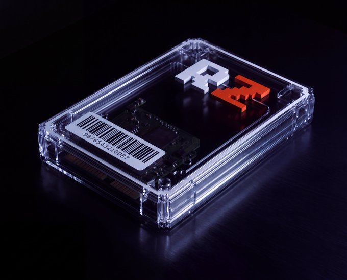 A clear acrylic case sits on a dark surface, housing an electronic component and labeled with a barcode reading '9876543210987' alongside white and red 3D 'R' letters.