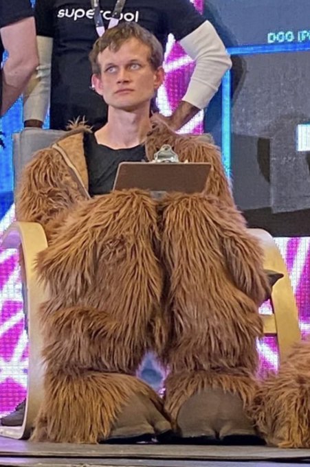A man sits in a large yellow chair wearing a full-body brown furry costume over a black shirt, holding a clipboard.