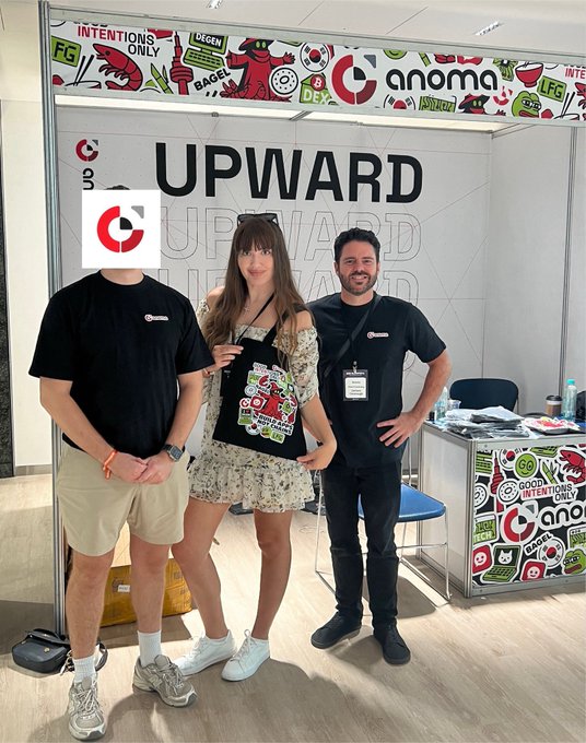 Three people pose at a trade show booth branded with 'UPWARD' and 'anoma' logos, with one person in the center holding a branded tote bag.