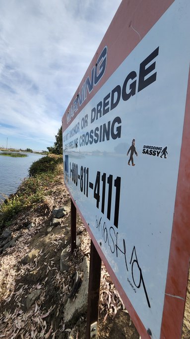 A warning sign reading 'WARNING DREDGE PIPELINE CROSSING' with a phone number '1-800-881-4111', located outdoors near a body of water and grass.